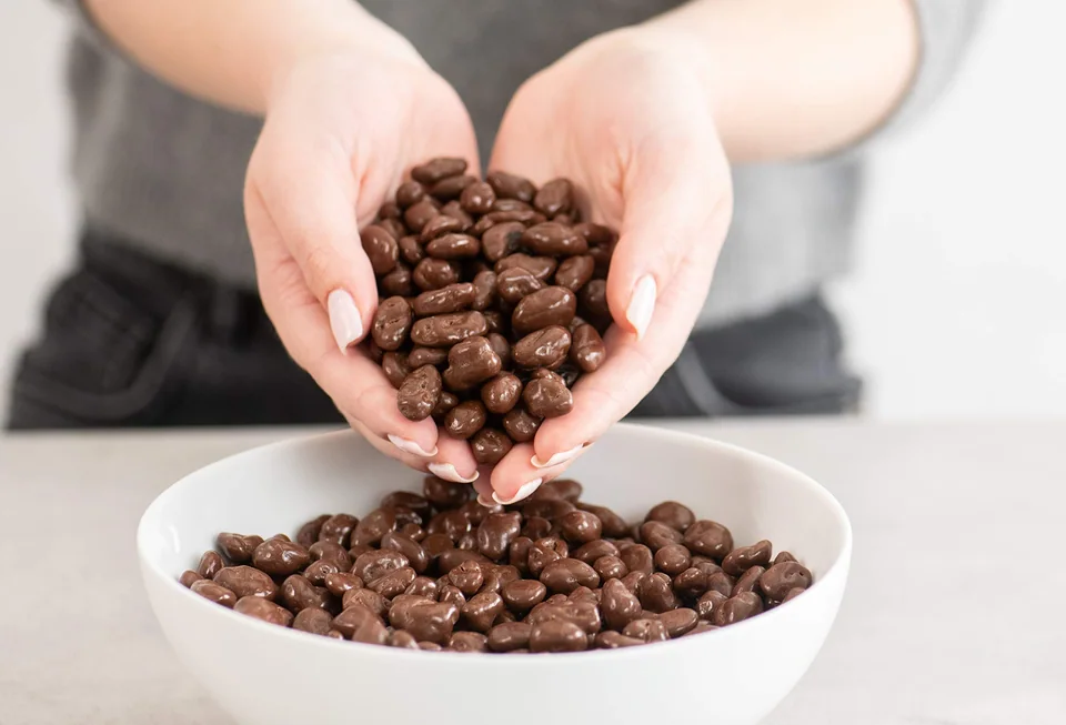 Two hands scooping chocolate-covered raisins over a white bowl